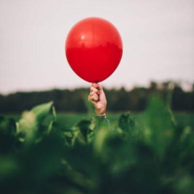 Hand holding a red ballon peaking over a green hedge